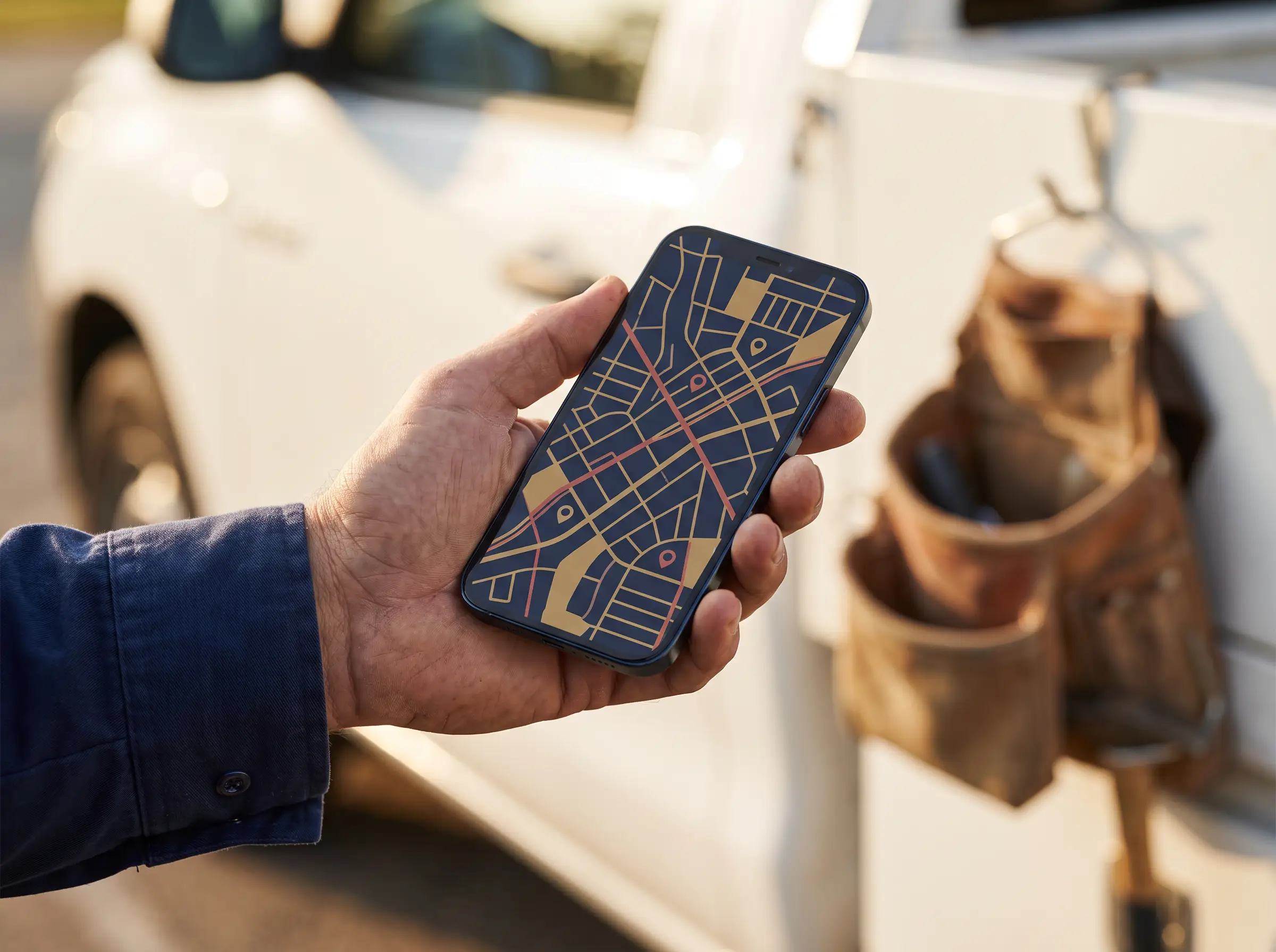 a tradie's hand holding a smartphone showing a map with location pins, work ute and tool belt in the background